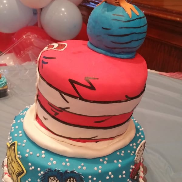 Colorful tiered birthday cake decorated with Dr. Seuss characters, red-and-white stripes, blue frosting, and balloons behind it.