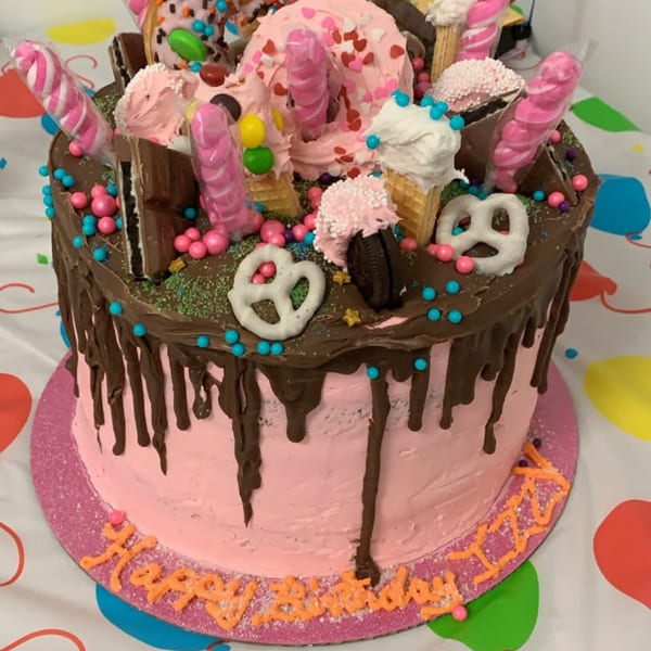 Elaborately decorated pink and chocolate drip cake topped with ice cream cones, candy, pretzels, sprinkles, and pink twisted lollipops.