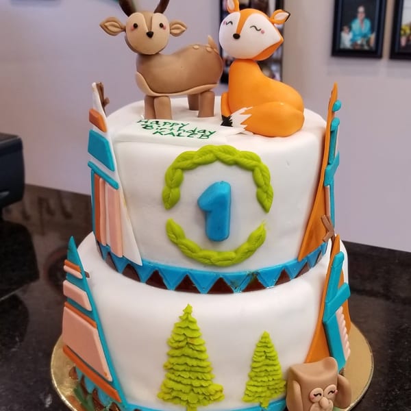 Two-tier woodland-themed birthday cake decorated with a deer, fox, trees, skis, and a blue number one.