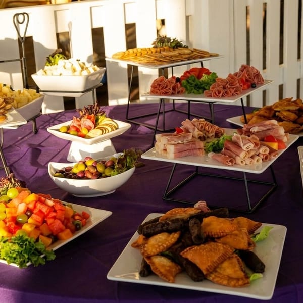 Buffet spread featuring assorted pastries, sliced meats, fruit, olives, fried appetizers, and vegetables on a purple tablecloth.