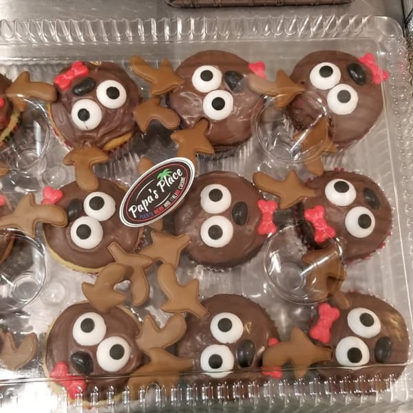 Tray of chocolate-covered cupcakes decorated as whimsical reindeer with candy eyes, antlers, noses, and red bows.