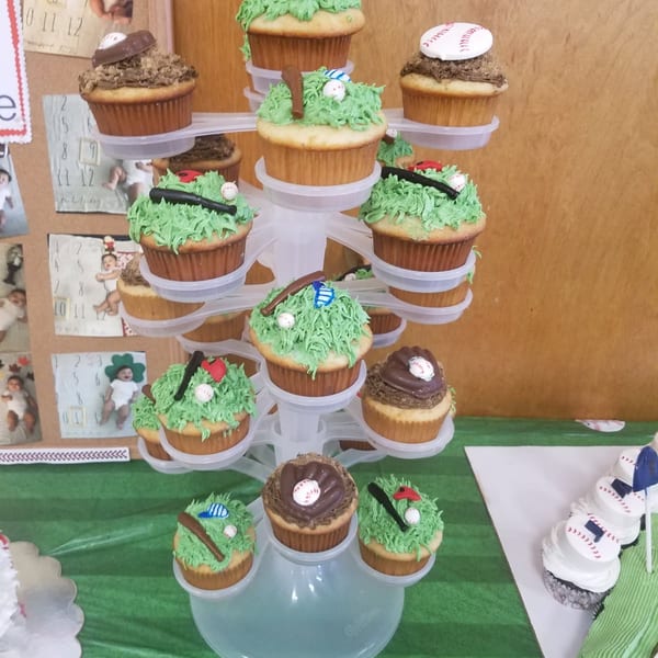 Decorated baseball-themed cupcakes arranged on a tiered stand, with green frosting, chocolate bats, baseballs, and red accents.