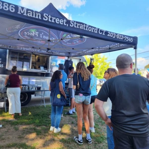 People wait at a colorful food truck beneath a canopy marked 386 Main Street, Stratford, Connecticut, on a sunny day.