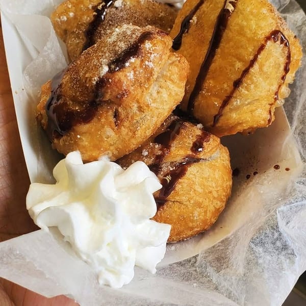 Golden fried pastries topped with powdered sugar, chocolate drizzle, and whipped cream, served in a paper-lined tray.