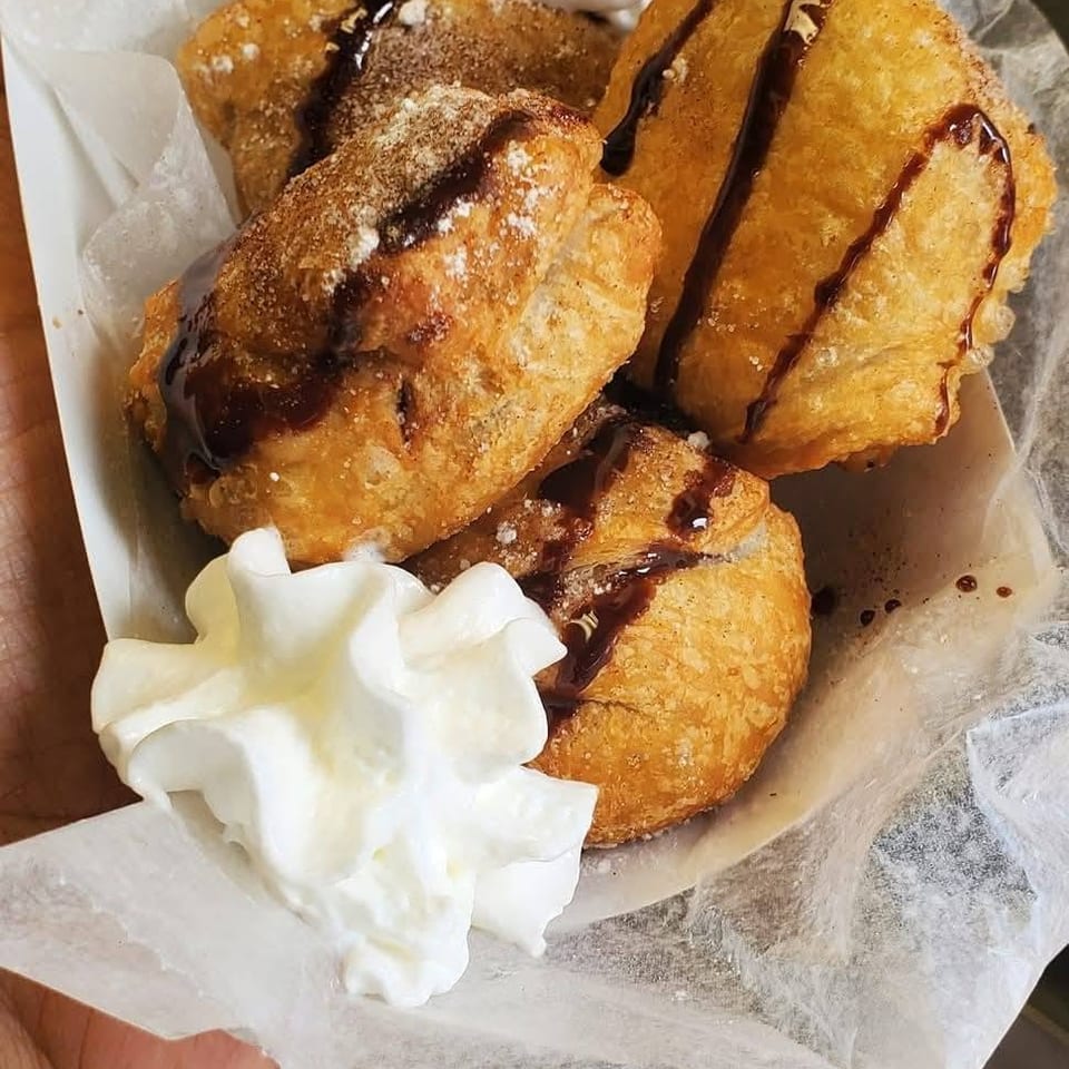 Golden fried pastries topped with powdered sugar, chocolate drizzle, and whipped cream, served in a paper-lined tray.