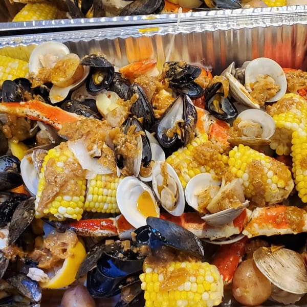 Seafood boil in aluminum trays with lobster, crab legs, shrimp, mussels, clams, corn, potatoes, and seasoned butter sauce.