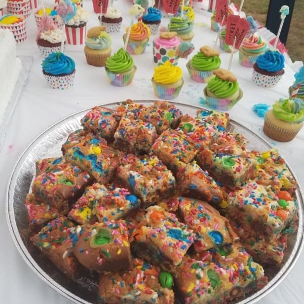 Colorful sprinkle-covered dessert bars on a platter, surrounded by brightly decorated cupcakes and candy-topped treats.