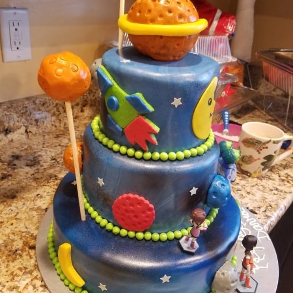 Three-tiered blue space-themed cake decorated with planets, rockets, stars, astronauts, and a Saturn-like topper.