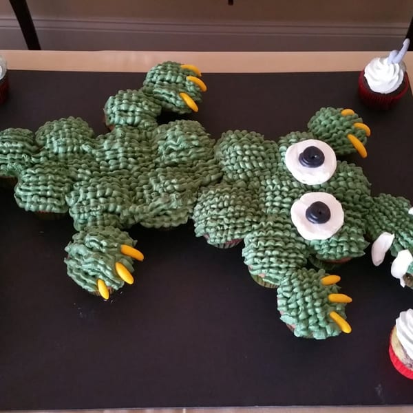 Large green crocodile-shaped cake decorated with candy spikes, eyes, claws, and surrounded by cupcakes on a black serving board.