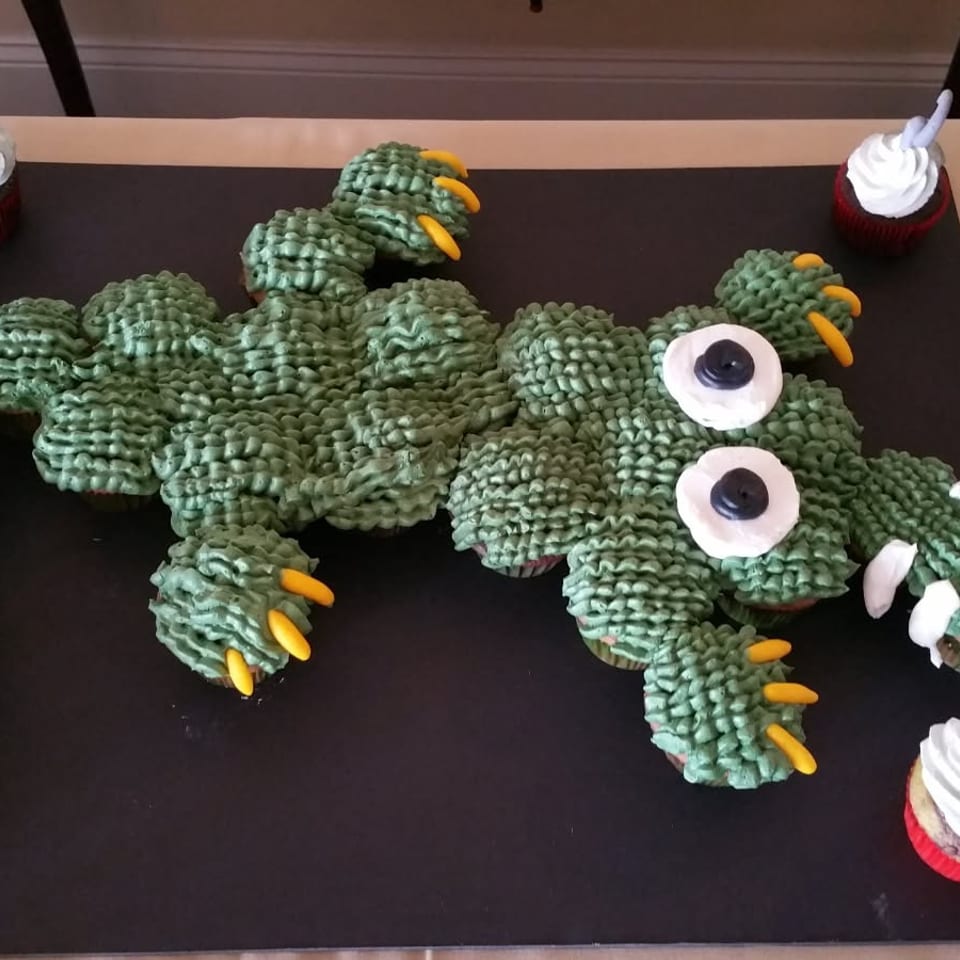 Large green crocodile-shaped cake decorated with candy spikes, eyes, claws, and surrounded by cupcakes on a black serving board.