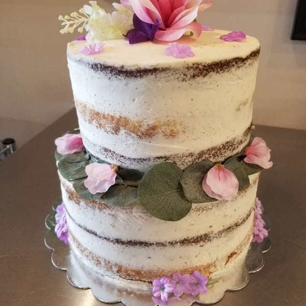 Two-tier naked cake decorated with pink flowers, greenery, and a large floral topper on a silver scalloped board.