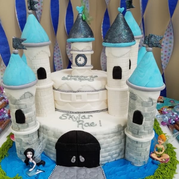 Castle-shaped birthday cake with turquoise roofs, glittering towers, a clock, “Skylar Rae” lettering, and mermaid fairy figurines.